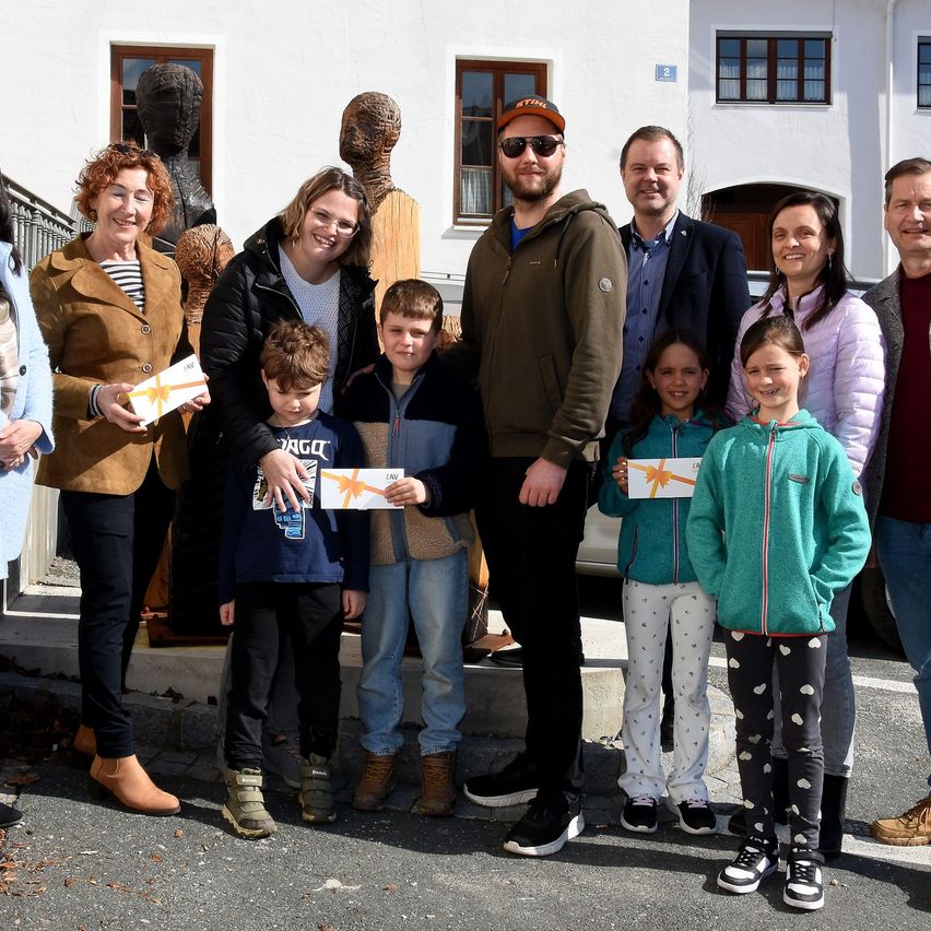 A group of adults and children stand outside a building, holding gift boxes. The adults are smiling, and some wear glasses. Two wooden sculptures are behind them.