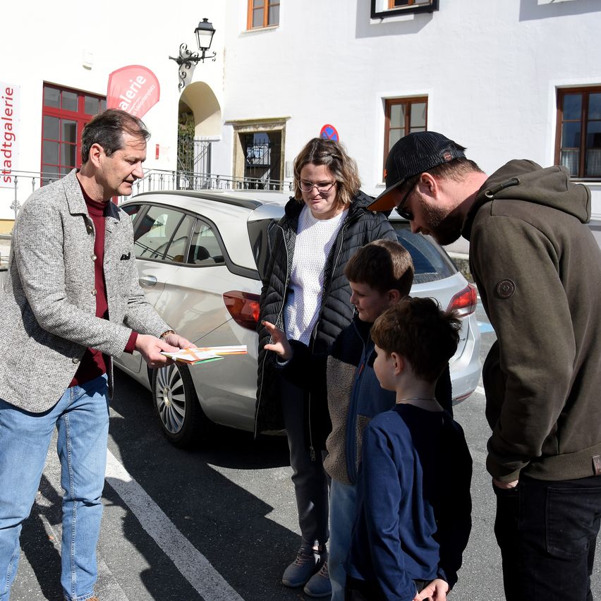A family of five stands in front of a silver car. The man and woman are talking to a boy holding a book. The man on the right wears a black cap and jacket. Behind them is a building with red windows and a banner.