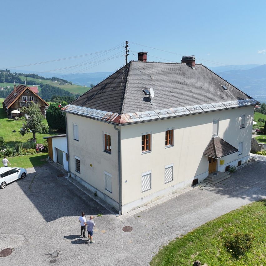 Eine Vogelperspektive eines weißen Hauses mit grauem Dach und Schornsteinen. Zwei Personen stehen davor, und ein weißes Auto ist geparkt. Im Hintergrund ein kleineres Haus und üppige Grünflächen.