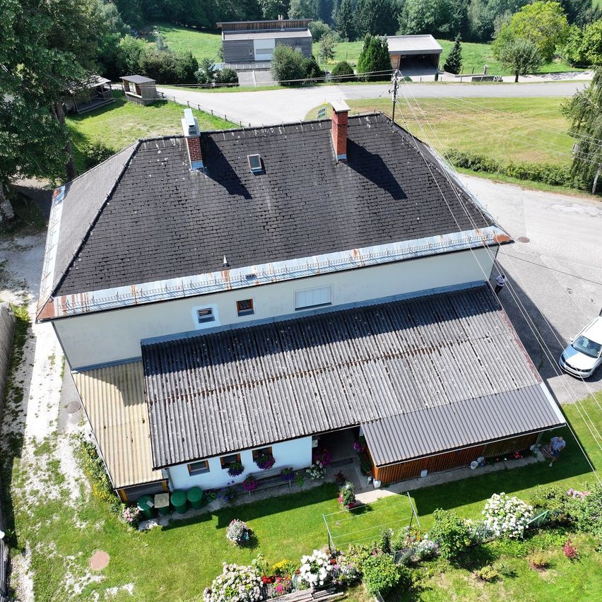 Ein großes weißes Haus mit schwarzem Dach und Schornsteinen ist von einem Garten mit Blumen und Bäumen umgeben. Ein Auto ist auf der Seite geparkt. In der Ferne befinden sich Gebäude und eine Straße.
