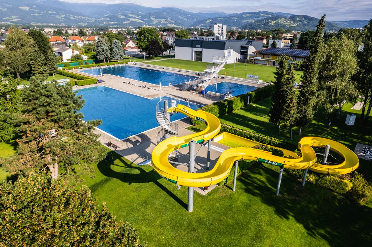 Eine Luftaufnahme eines Schwimmbeckens mit einer gelben Rutsche und einer weißen Leiter. Bäume und Gebäude im Hintergrund.