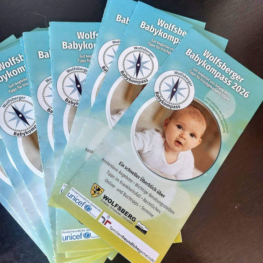 Ein Stapel farbenfroher Broschüren mit dem Titel 'Babykompass 2026', die ein Bild eines Babys und einen Kompass zeigen, liegt auf einer dunklen Oberfläche. Die Broschüren bieten Eltern Tipps und werden von UNICEF und Familienfreundliche Gemeinden gesponsert.