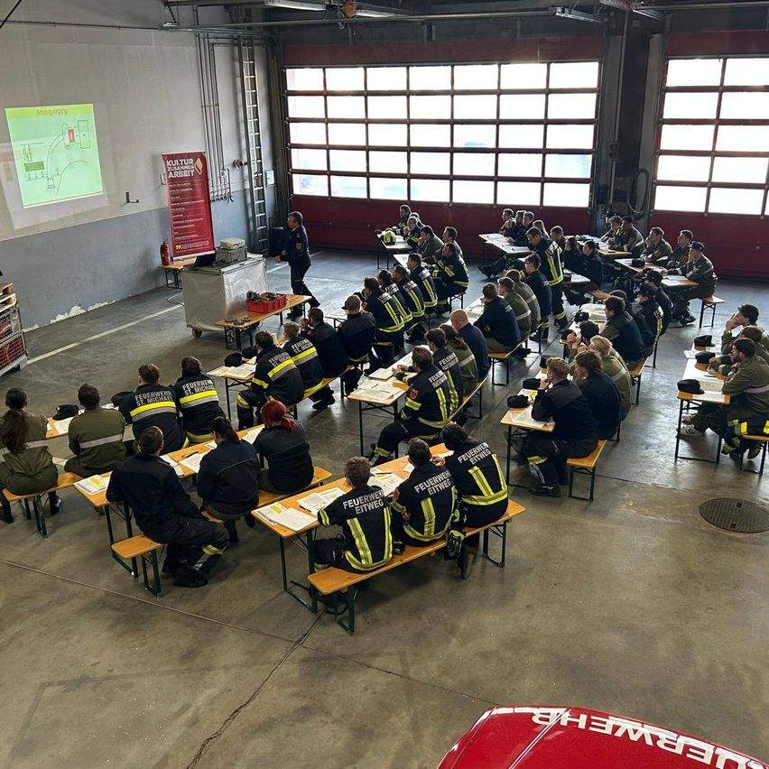 Feuerwehrleute nehmen an einer Schulung in einem großen Raum mit großen Fenstern teil. Sie sitzen an Tischen mit Papieren und tragen Uniformen. Eine Projektionsleinwand zeigt ein Diagramm.