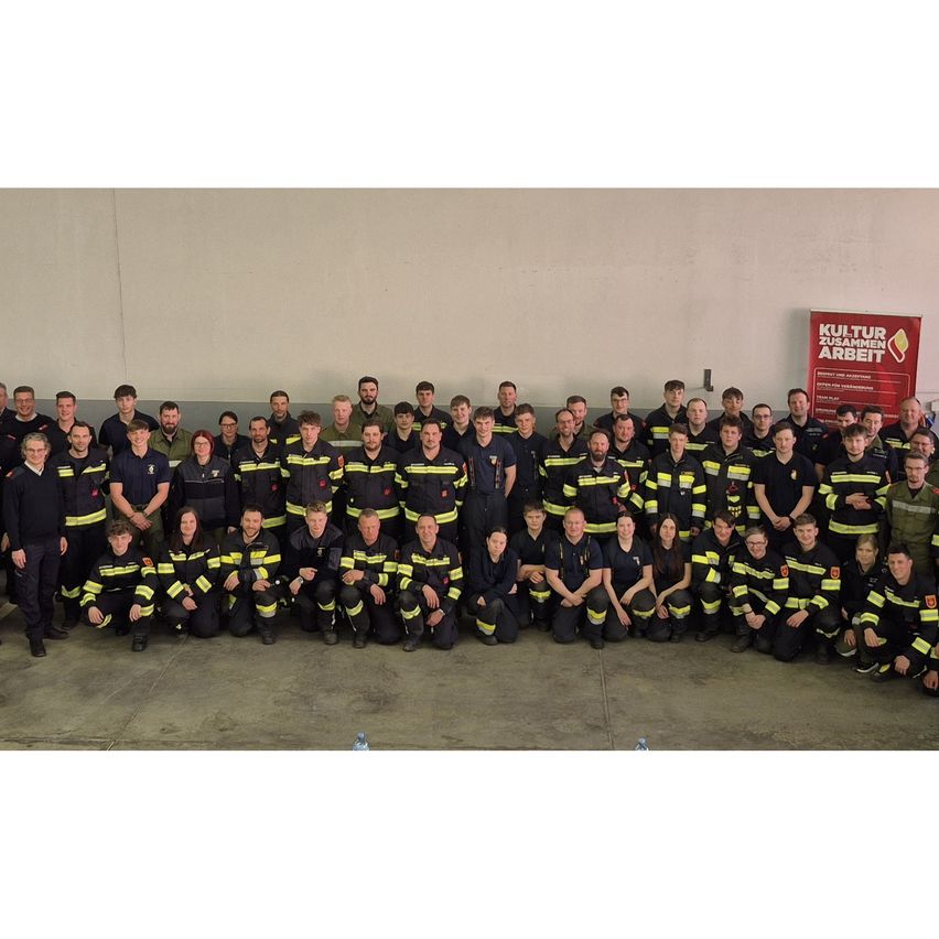Eine große Gruppe von Feuerwehrleuten in Uniform, einige kniend, posiert für ein Foto in einem Raum. Ein Banner mit der Aufschrift Kultur Zusammen Arbeiten befindet sich rechts.