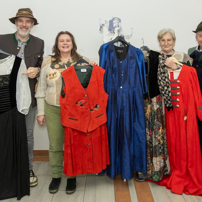 Eine Gruppe von Menschen posiert für ein Foto und hält traditionelle Kleidung. Sie stehen vor einer weißen Wand.