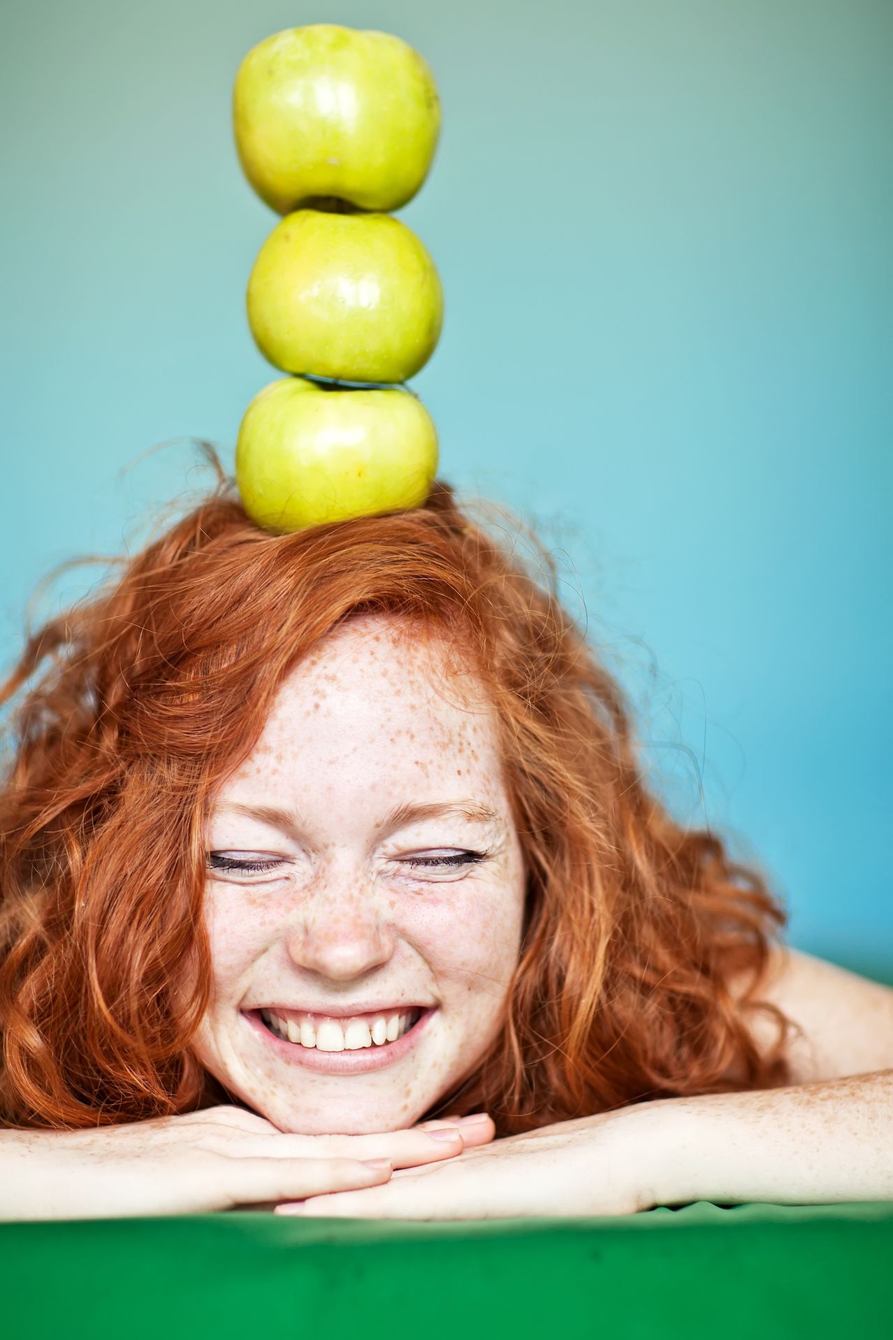 Eine Frau mit roten Haaren und Sommersprossen lächelt, während zwei grüne Äpfel auf ihrem Kopf balancieren.