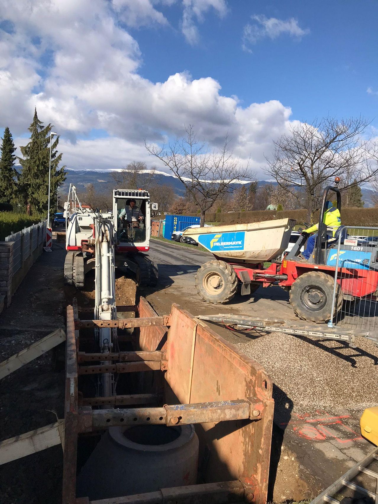 Zwei Baufahrzeuge stehen auf einer Straße, eines gräbt und das andere ist mit einem Eimer beladen. In der Nähe befinden sich Bäume, Büsche und ein Zaun, mit Bergen im Hintergrund.