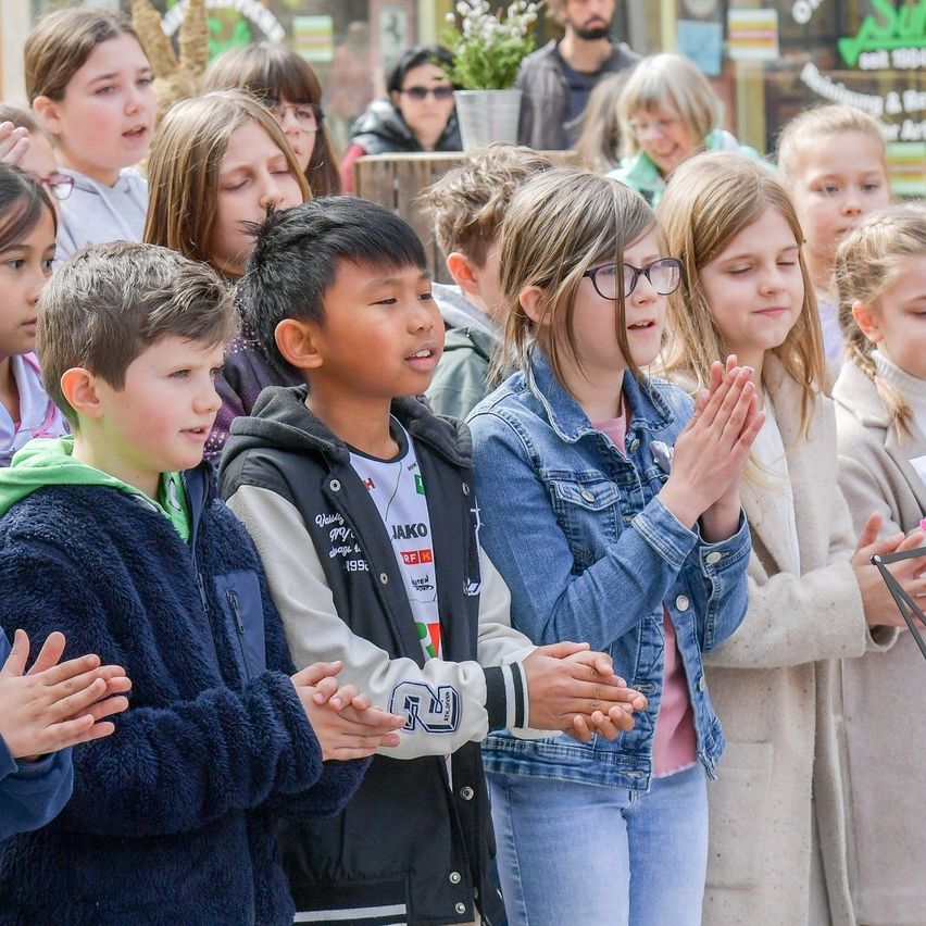 Kinder stehen in einer Reihe, klatschen und lächeln, wahrscheinlich in einem Klassenzimmer oder einer Gemeinschaftsveranstaltung. Ein Mädchen in Jeans und ein Junge in einer grünen Kapuzenjacke sind vorne.
