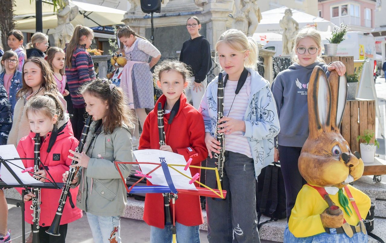 Vier Mädchen spielen Klarinetten im Freien. Das Mädchen im roten Mantel hält einen Notenständer. Hinter ihnen ist eine große Holzskulptur zu sehen.