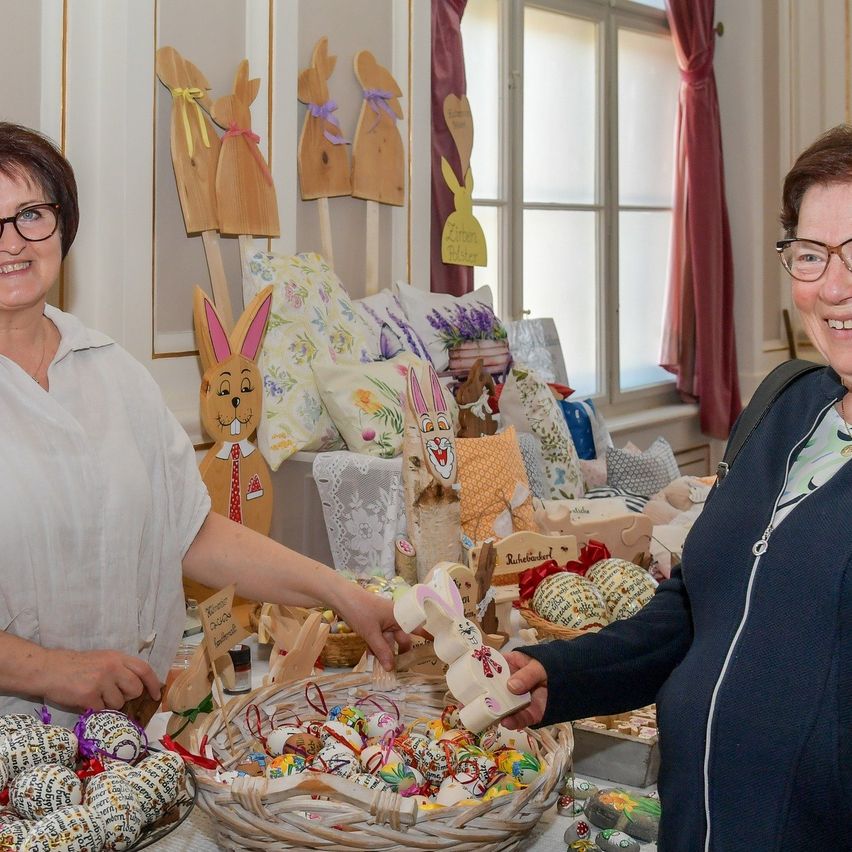 Zwei Frauen lächeln und stehen neben einem Tisch voller Osterdekoration und Eiern. Eine hält eine Hasenfigur und die andere einen Korb mit Eiern. Dahinter sind hölzerne Hasen und Kissen ausgestellt.