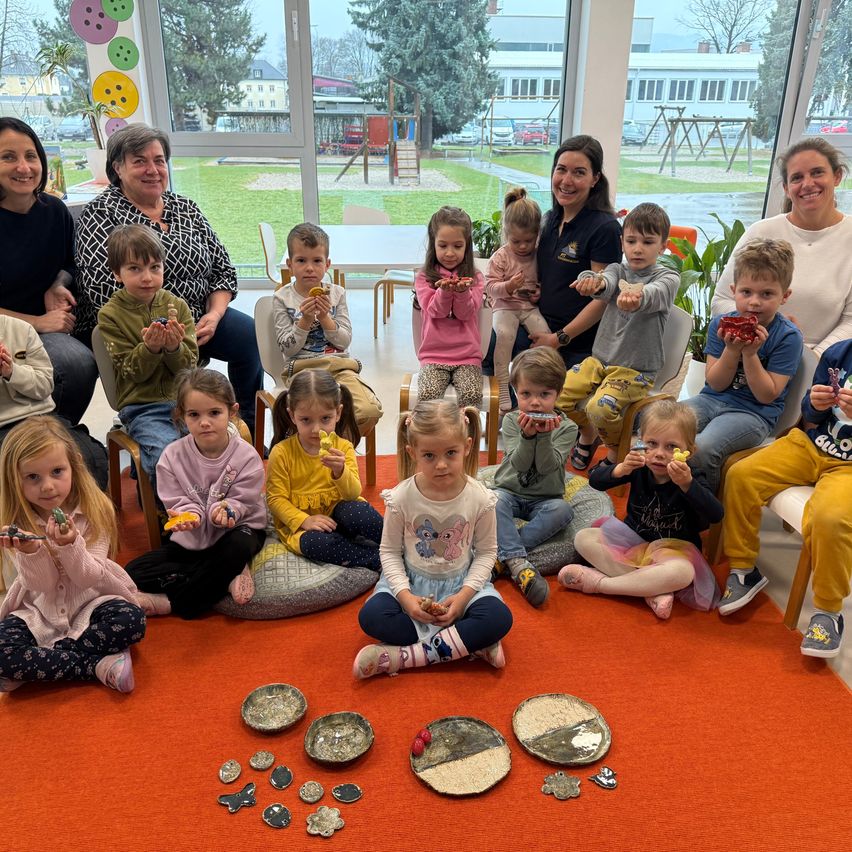 Eine Gruppe von Kindern und Erwachsenen sitzt in einem Kreis auf einem roten Teppich und hält Gegenstände aus Ton. Sie scheinen an einer Kunstaktivität teilzunehmen. Einige lächeln. Der Hintergrund zeigt einen Raum mit Glaswänden und einem Spielplatz.