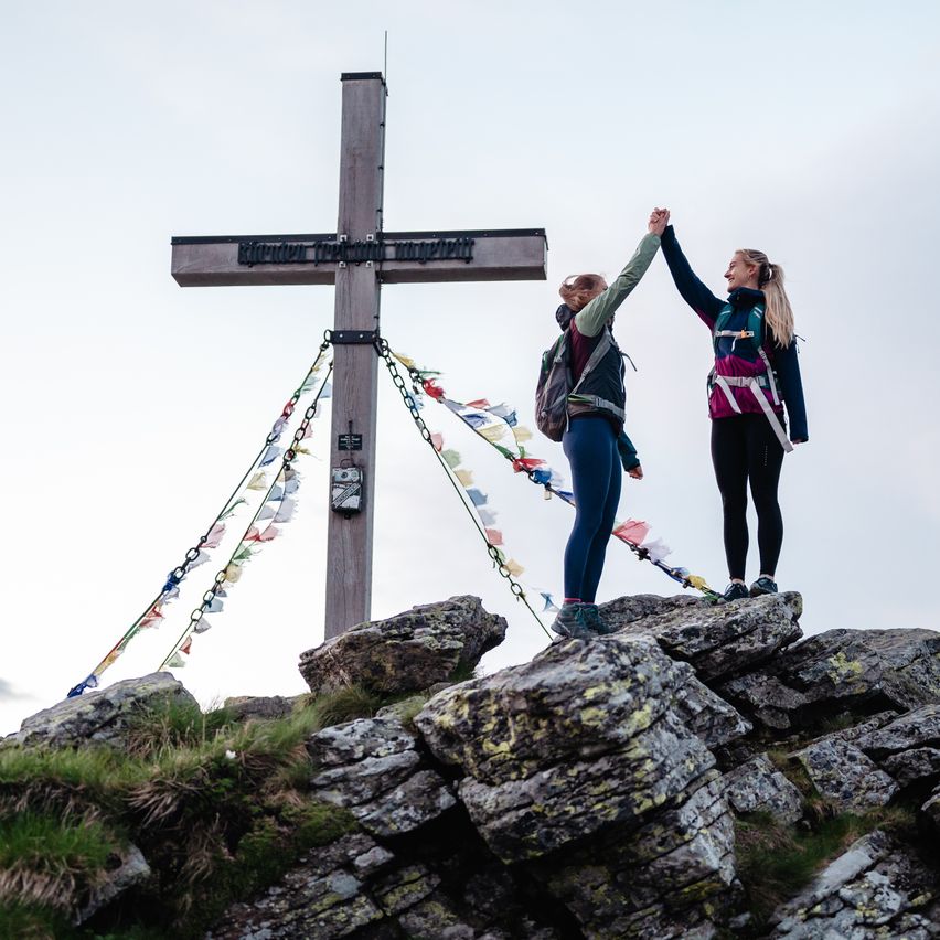 Two women stand on a rocky mountain peak, high-fiving each other in front of a wooden cross adorned with colorful prayer flags.