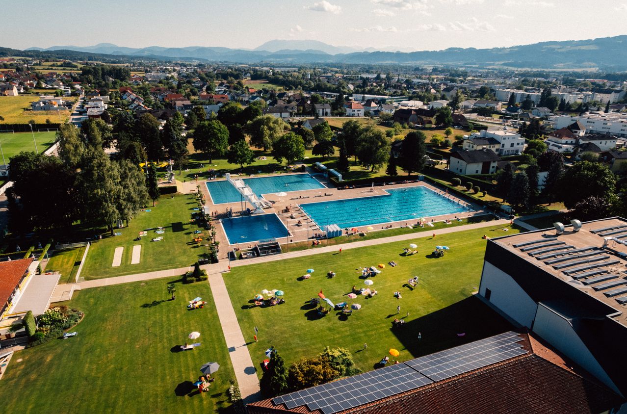 An aerial view of a large outdoor swimming pool with people sunbathing and swimming, surrounded by a green park with trees and buildings in the background.