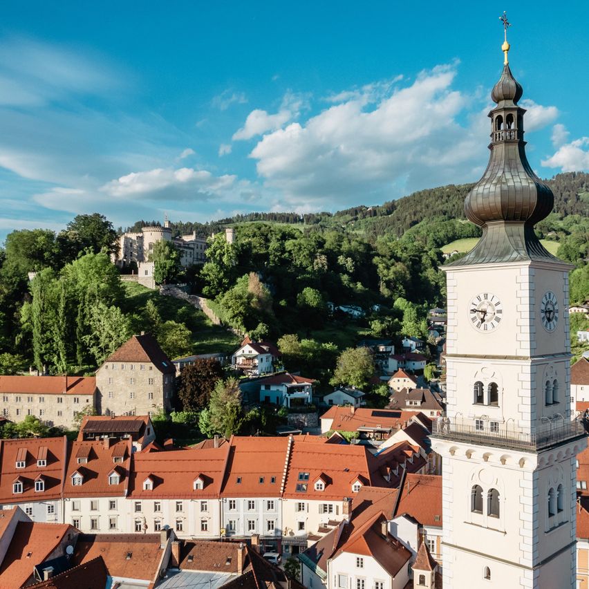Aerial view of a town with red roofs, a tall clock tower, and a castle in the distance under a blue sky with clouds.