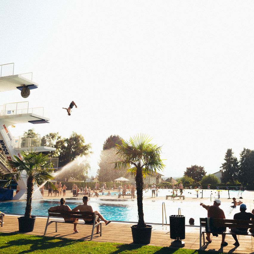 A person dives into a large outdoor pool while others relax on benches around the pool. Trees and a diving tower are in the background.
