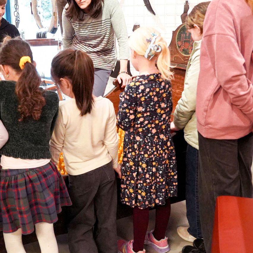A group of young girls and adults are gathered around a table, possibly in a museum or educational setting. The girls are wearing various outfits, and a woman in a striped shirt is standing behind them.