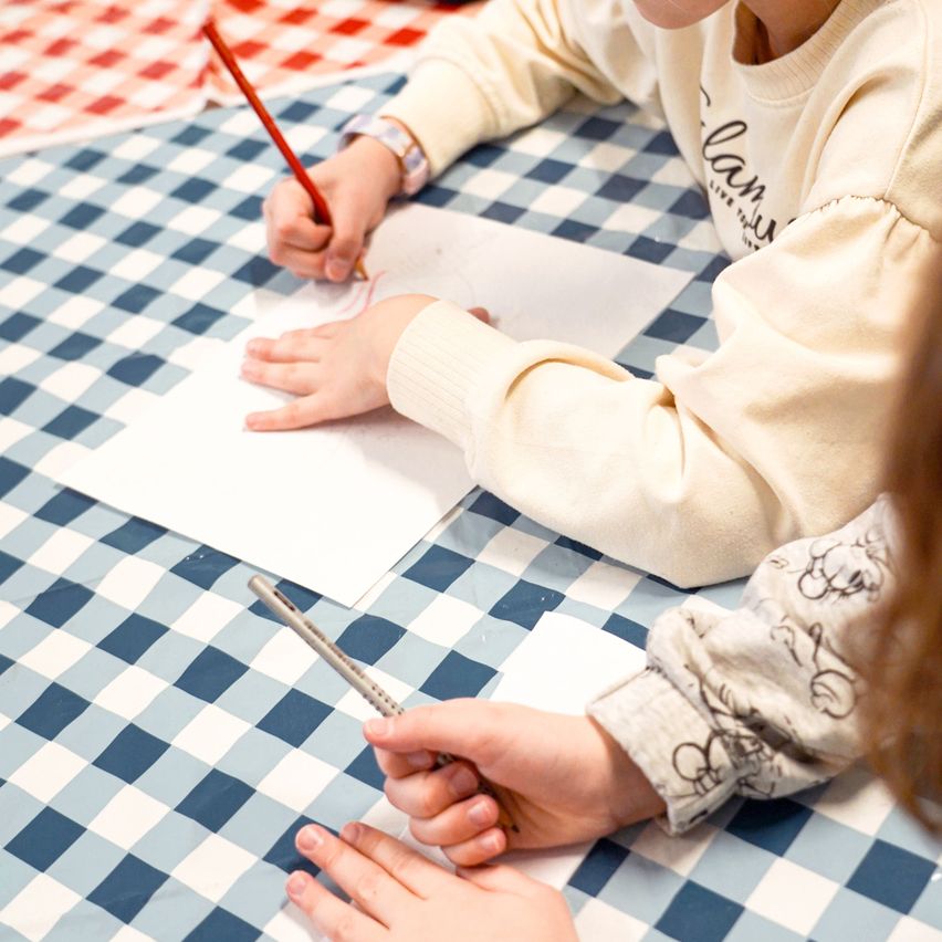 A child is sitting at a table, drawing on a piece of paper with a pencil. The table is covered with a blue and white checkered tablecloth.