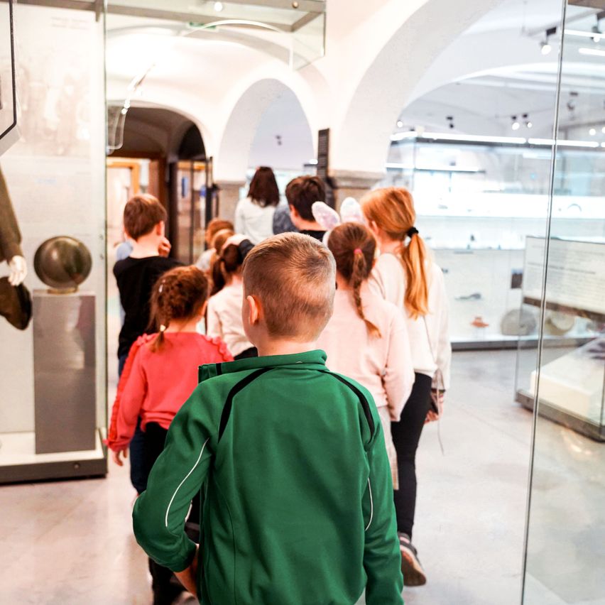 A group of children are walking through a museum, with a boy in a green jacket leading the way. Behind him, a girl in a pink shirt and another in a black shirt follow closely. The museum is filled with glass displays and exhibits.