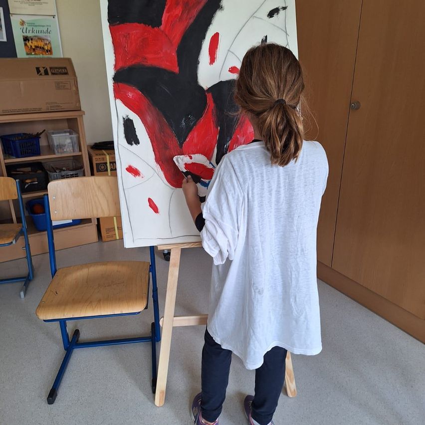 A young girl is painting a large canvas with red and black paint on an easel in a room.