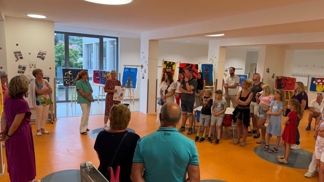 A group of people are gathered in a room with paintings on easels. Some are standing, while others are sitting on chairs. A woman in a green shirt is holding a book.