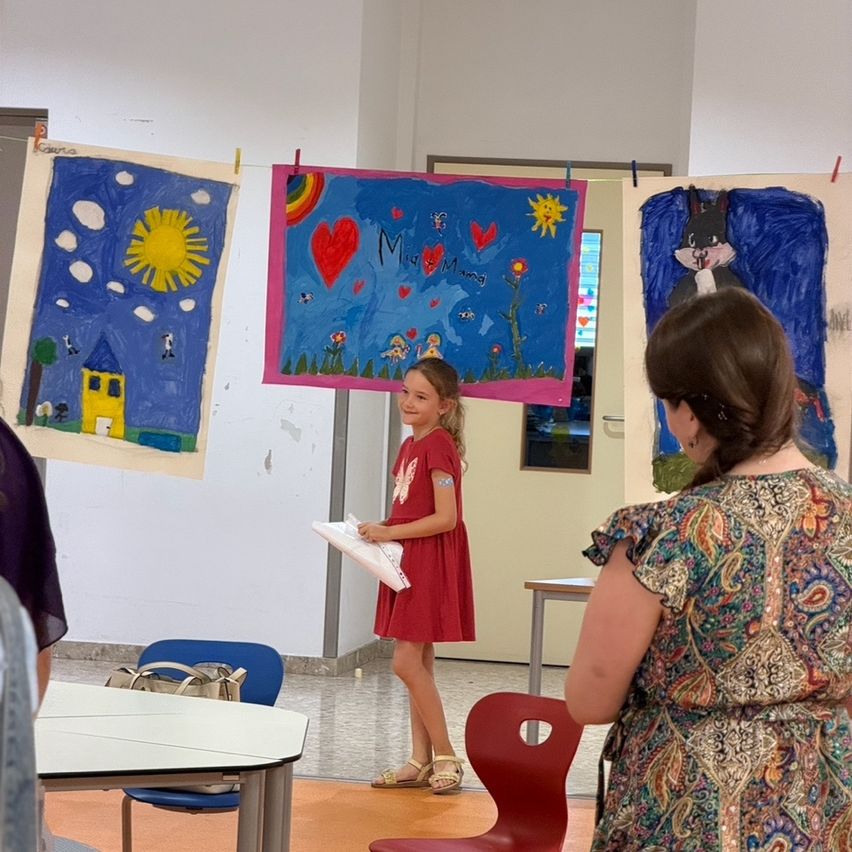 A young girl in a red dress stands in front of a classroom wall with various colorful paintings. She holds a piece of paper and looks towards an adult woman.