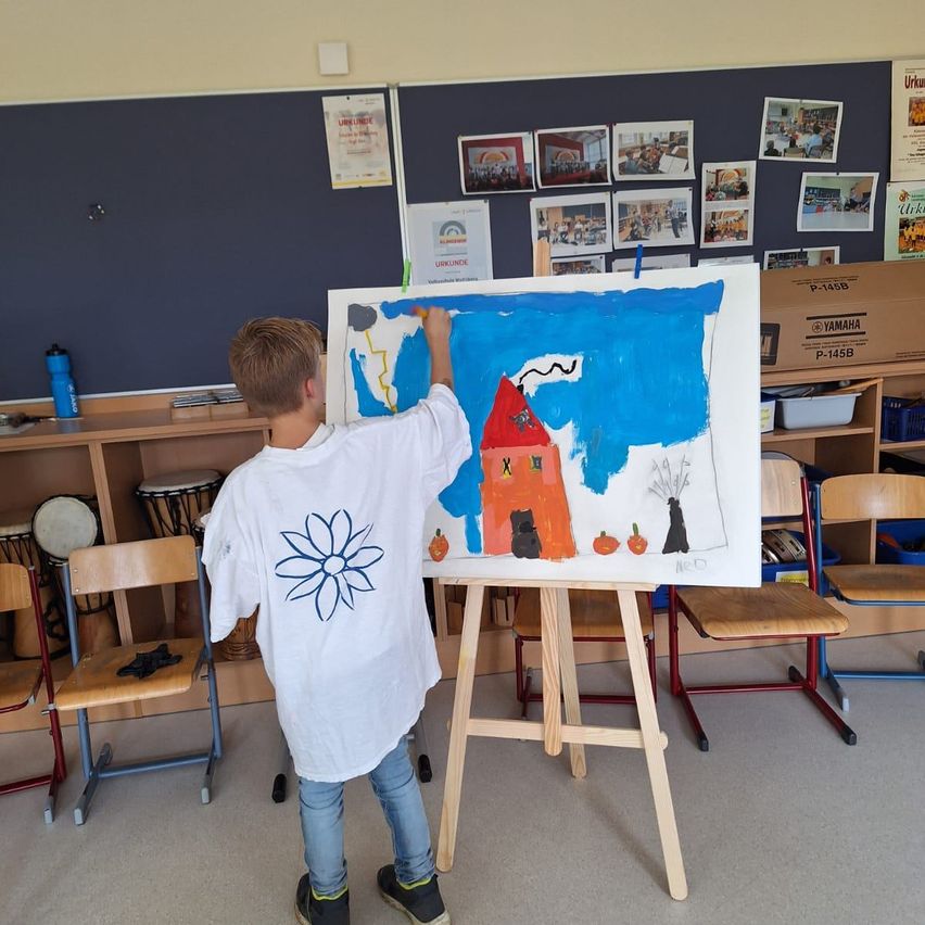 A young boy stands at an easel, painting a picture of a house with a red roof and pumpkins in front. He is in a classroom with various pictures on the wall and chairs around him.