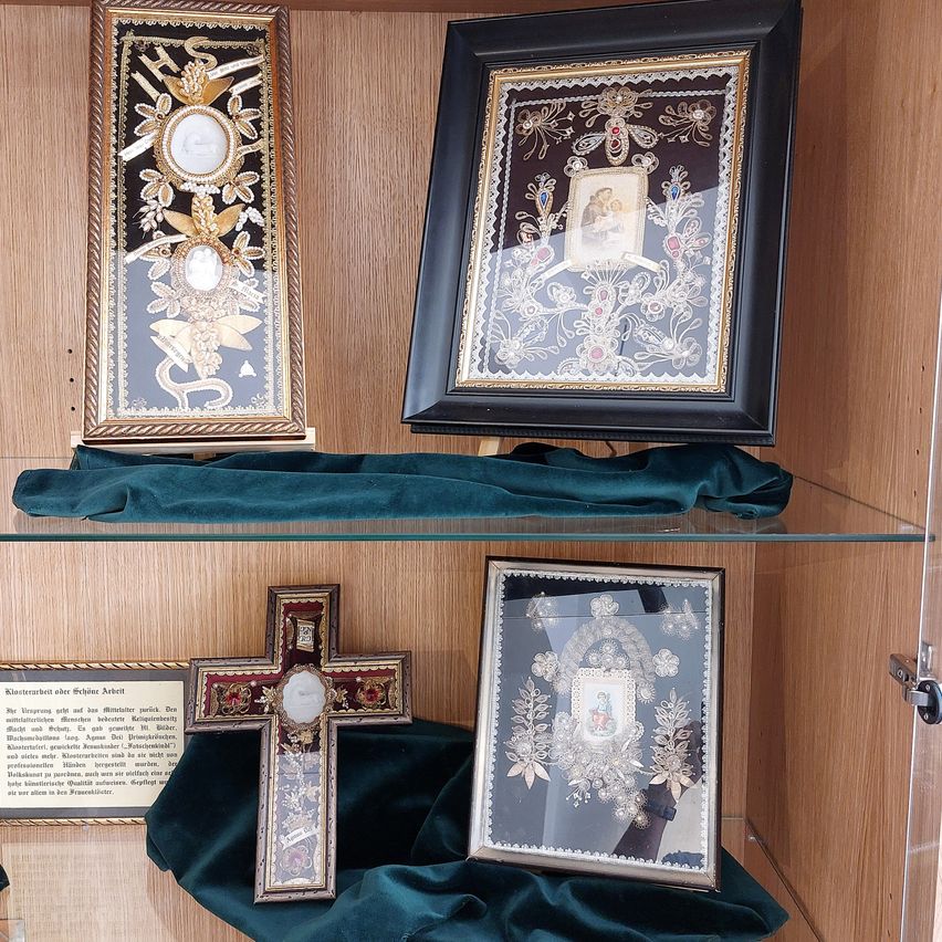A wooden shelf displays three ornate framed pieces, including a cross and two portraits, one with a mother and child, all set against a dark background.