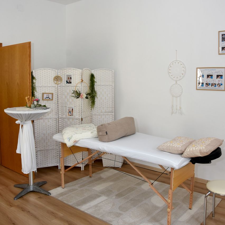 A massage table is placed on a rug in a room with white walls. A white table with a white cloth is on the left. A white screen is behind the table. A dream catcher is hanging on the wall.