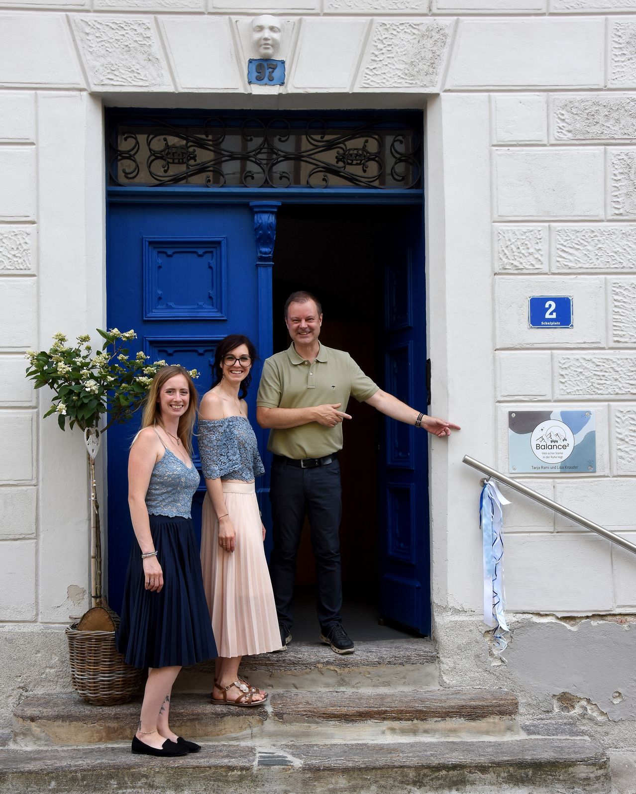 Three people stand in front of a blue door with a decorative plant, smiling and posing for a photo. The man points at the door.