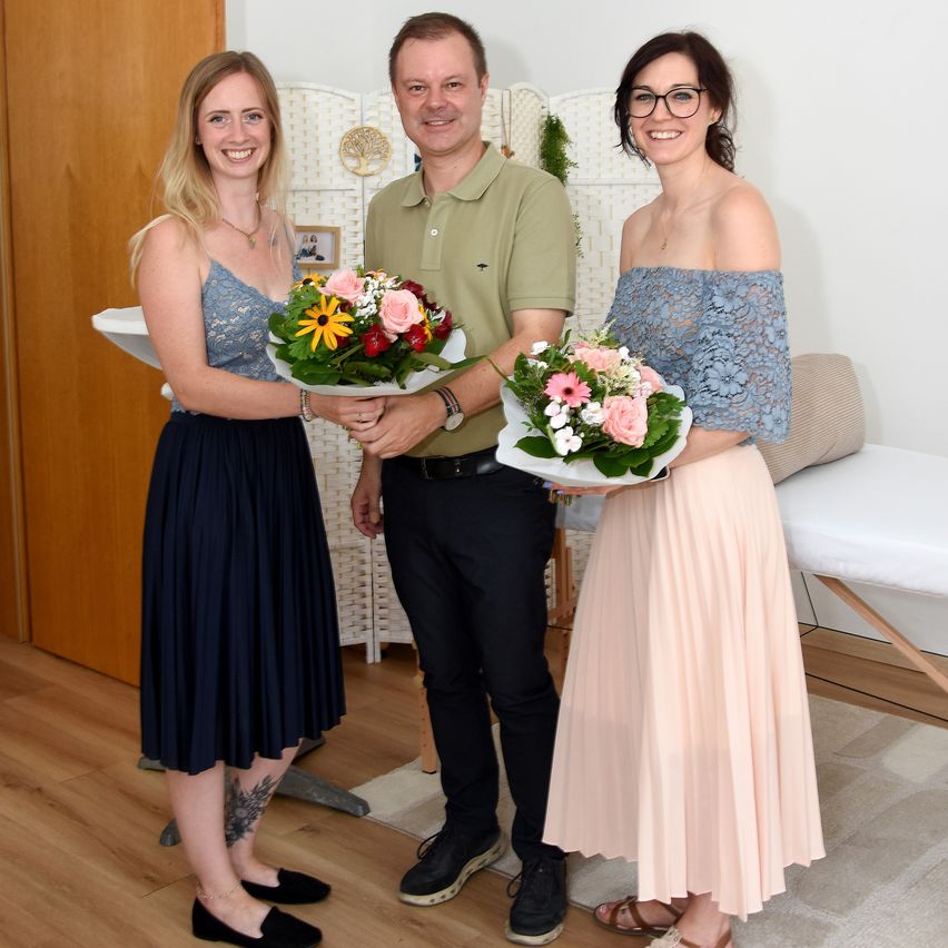 Three people stand together in a room, holding bouquets of flowers. A man stands in the middle, while two women stand on either side. The woman on the left wears a blue dress and black shoes, and the woman on the right wears a blue off-shoulder top and a pink skirt.