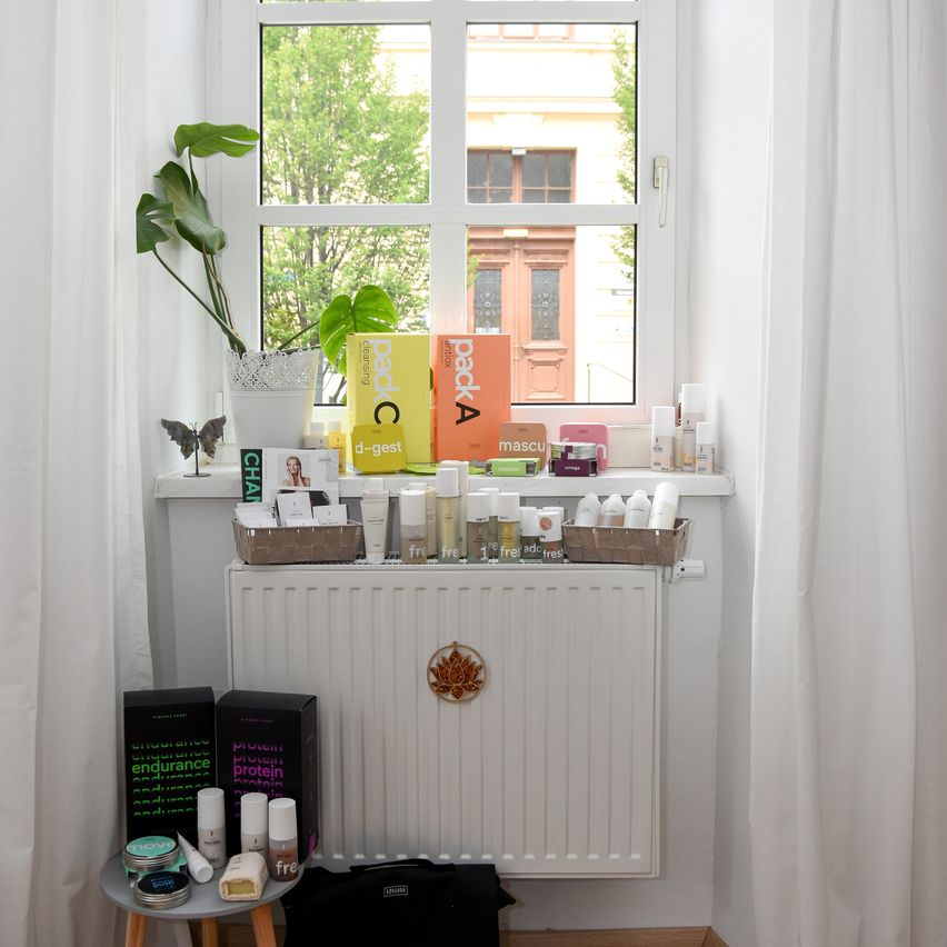 A white shelf with various products and a plant on a windowsill. The products include boxes, bottles, and a bag. A radiator is below the shelf.