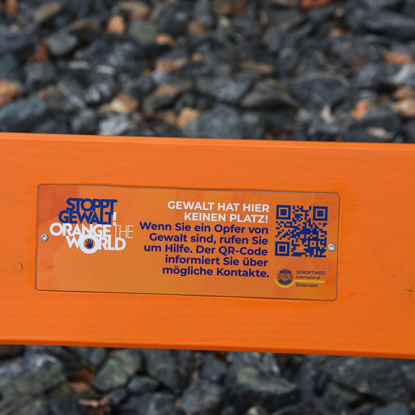 Ein orangefarbenes Schild mit dem Text 'STOPP GEWALT! ORANGE THE WORLD' und einem QR-Code. Das Schild ist auf einem orangefarbenen Metallstab über einem Kieshintergrund angebracht.