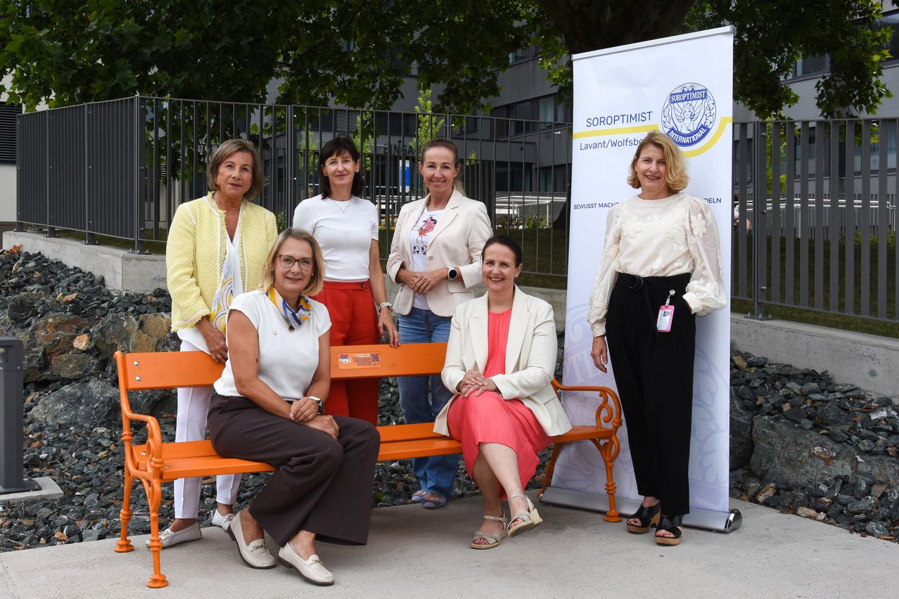 Fünf Frauen stehen hinter einer orangefarbenen Bank und lächeln für ein Foto. Sie tragen verschiedene Outfits, und ein Banner mit 'Soroptimist' ist hinter ihnen.