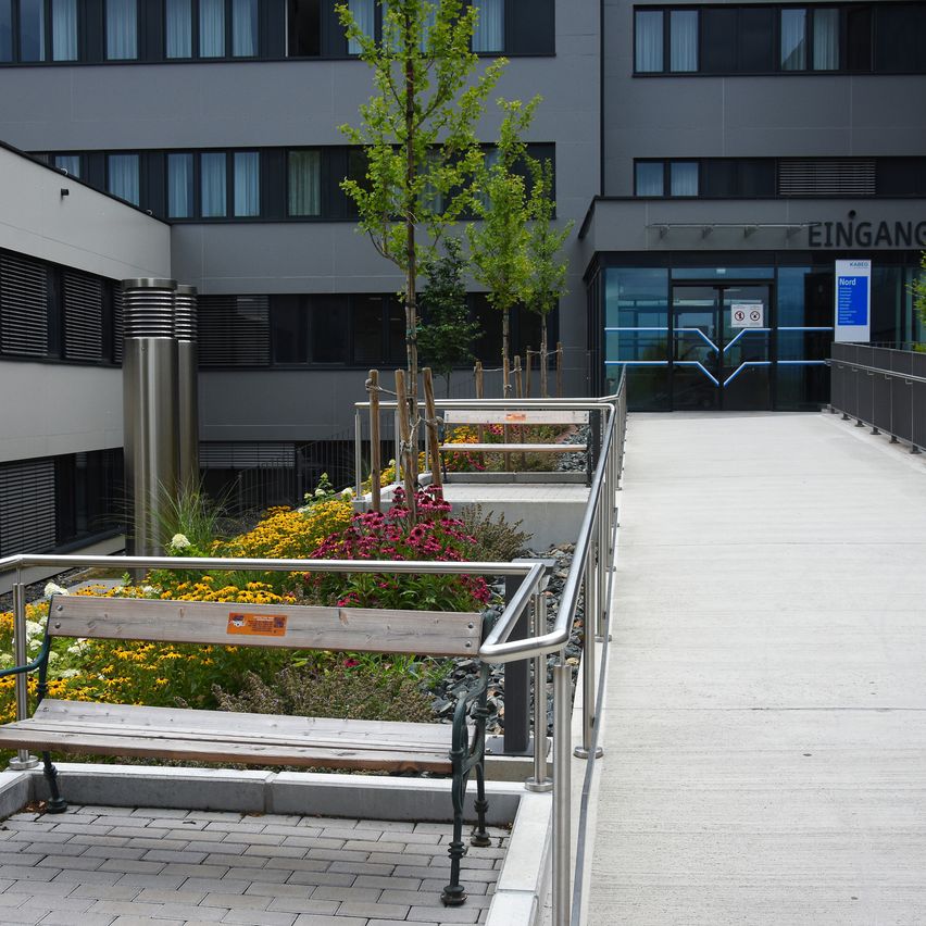 Ein modernes Gebäude mit einem Gartenbereich und einer Bank in der Nähe des Eingangs. Der Garten hat bunte Blumen und Bäume.
