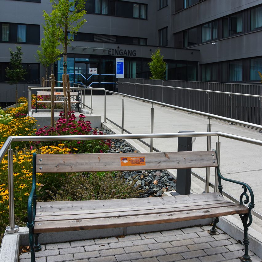 Eine Holzbank mit Metallarmlehnen steht auf einem Ziegelweg vor einem Gebäude mit dem Schild 'Eingang'. Die Bank ist von einem Garten mit bunten Blumen und Pflanzen umgeben.
