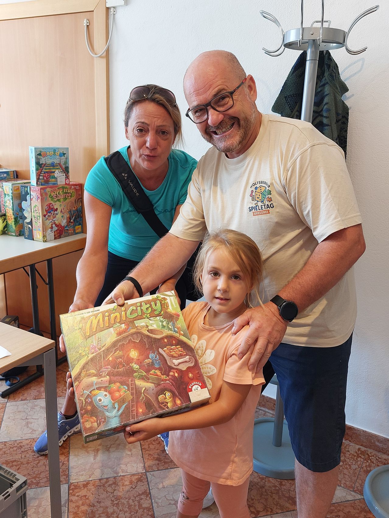 Ein Mann, eine Frau und ein junges Mädchen halten eine Brettspielbox, lächeln und posieren für ein Foto in einem Raum.