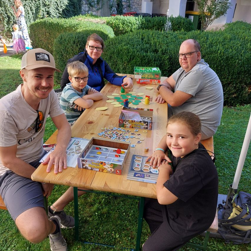 Eine vierköpfige Familie sitzt an einem Holztisch und spielt an einem sonnigen Tag in einem Garten ein Brettspiel.
