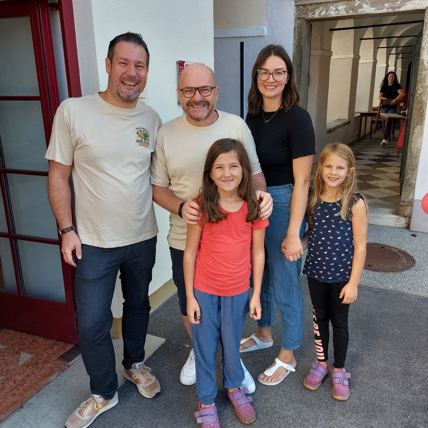 Eine vierköpfige Familie steht lächelnd für ein Foto vor einem Gebäude. Der Mann links trägt eine Brille und eine Uhr, während der Mann in der Mitte einen kahlen Kopf hat. Die Frau rechts trägt eine Brille und eine Halskette. Das Mädchen in der Mitte trägt ein rotes Shirt und blaue Hosen.