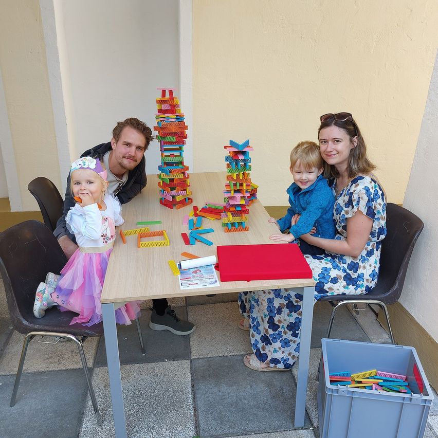 Eine Familie sitzt um einen Tisch mit bunten Bauklötzen, zwei Kinder spielen damit und eine Frau hält einen Kleinkind.