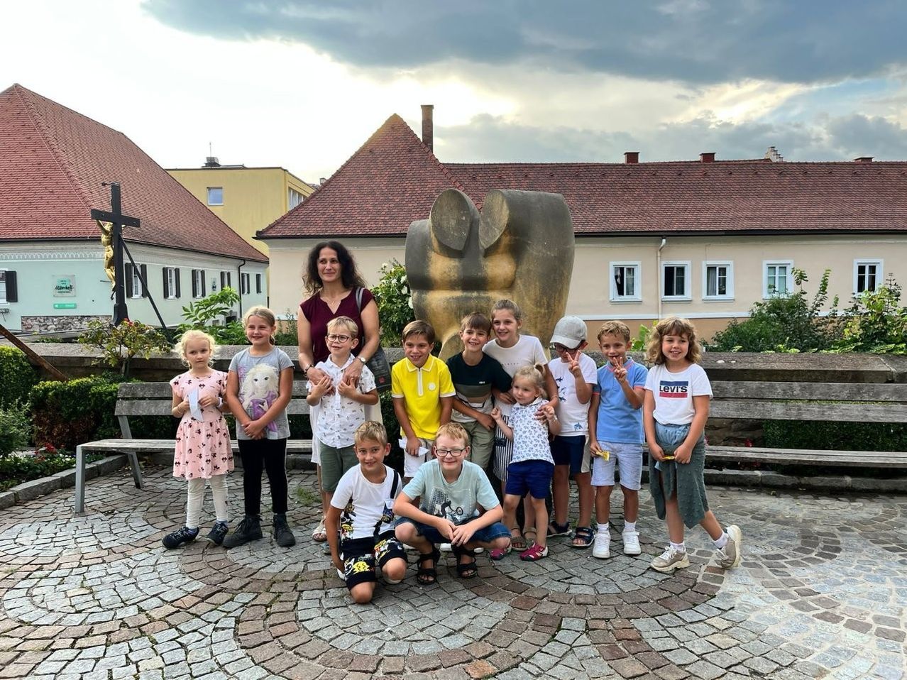 Eine Gruppe von Kindern und zwei Erwachsenen posiert für ein Foto vor einer großen Steinskulptur und einem Gebäude mit rotem Dach.
