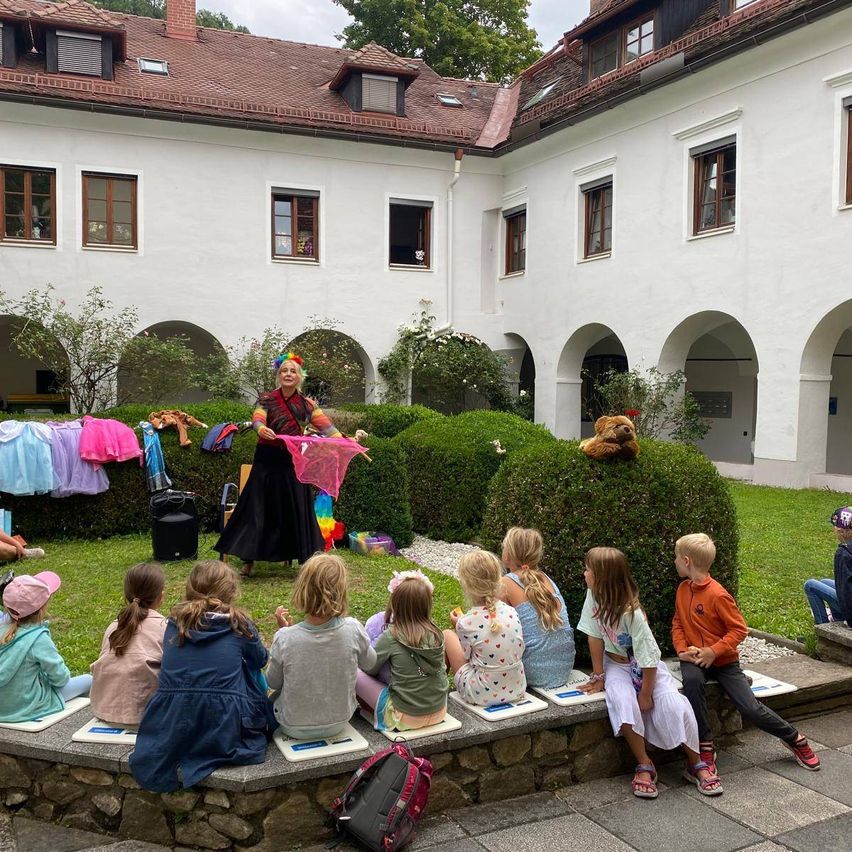 Eine Frau in bunter Kleidung führt vor einer Gruppe von Kindern auf, die auf einer Steinbank im Innenhof sitzen.