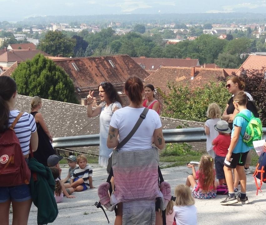 Eine Gruppe von Menschen, einschließlich Erwachsener und Kindern, versammelt sich auf einem Hügel mit Blick auf eine Stadt. Einige stehen, während andere auf dem Boden sitzen. Die Erwachsenen führen ein Gespräch.