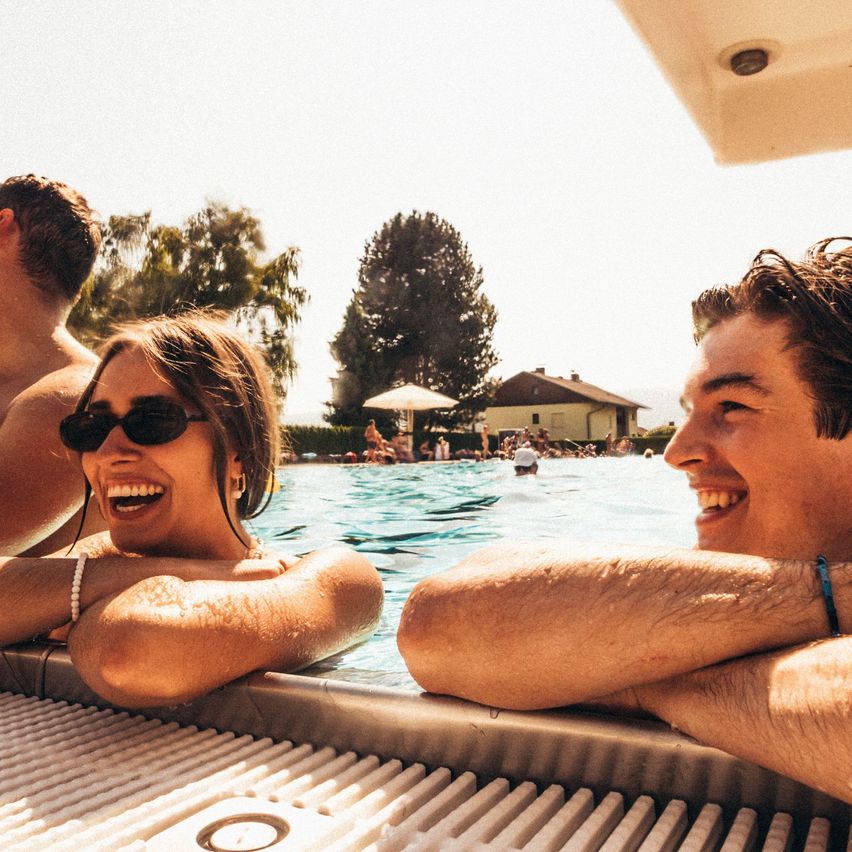 Eine Gruppe von Menschen genießt einen sonnigen Tag am Pool, wobei eine Frau mit Sonnenbrille und ein lächelnder Mann neben ihr zu sehen sind. Bäume und ein Haus sind im Hintergrund erkennbar.