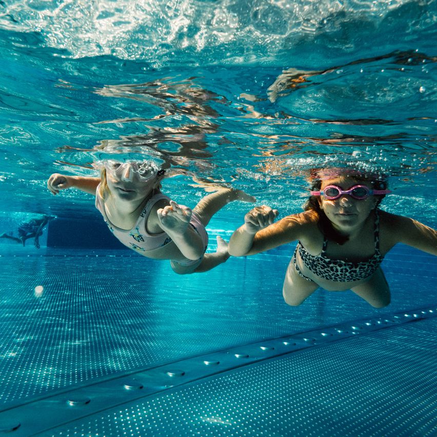 Zwei junge Mädchen mit Schwimmbrillen schwimmen unter Wasser in einem Pool, eine mit Schnorchel und die andere mit einem Leopardenprint-Badeanzug.
