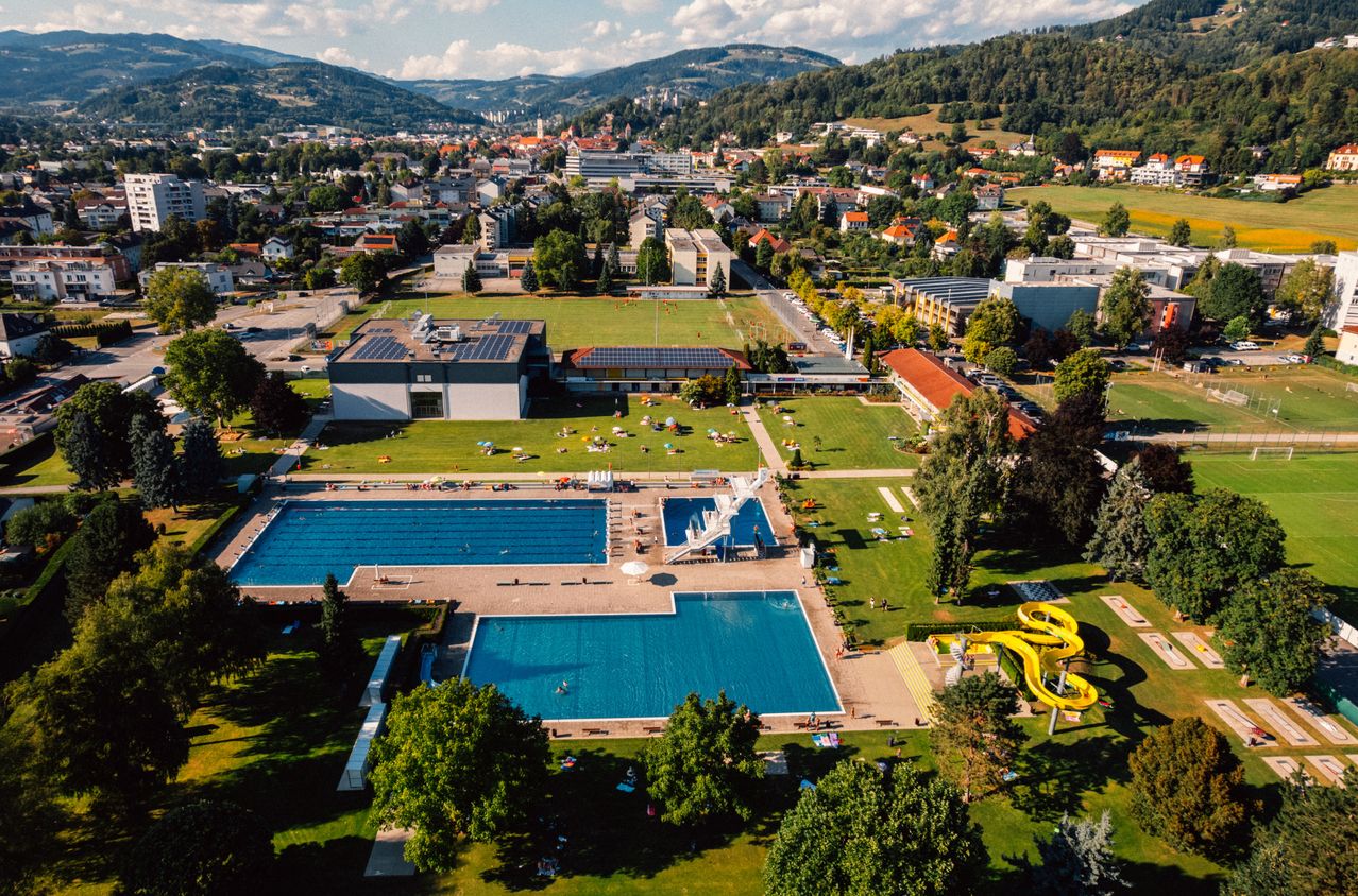 Eine Luftaufnahme eines großen Freibadkomplexes mit mehreren Becken, einer Sprungturm und einer Wasserrutsche. Das Gelände ist von saftigem grünem Gras und Bäumen umgeben, mit Gebäuden und einer Stadtlandschaft im Hintergrund.