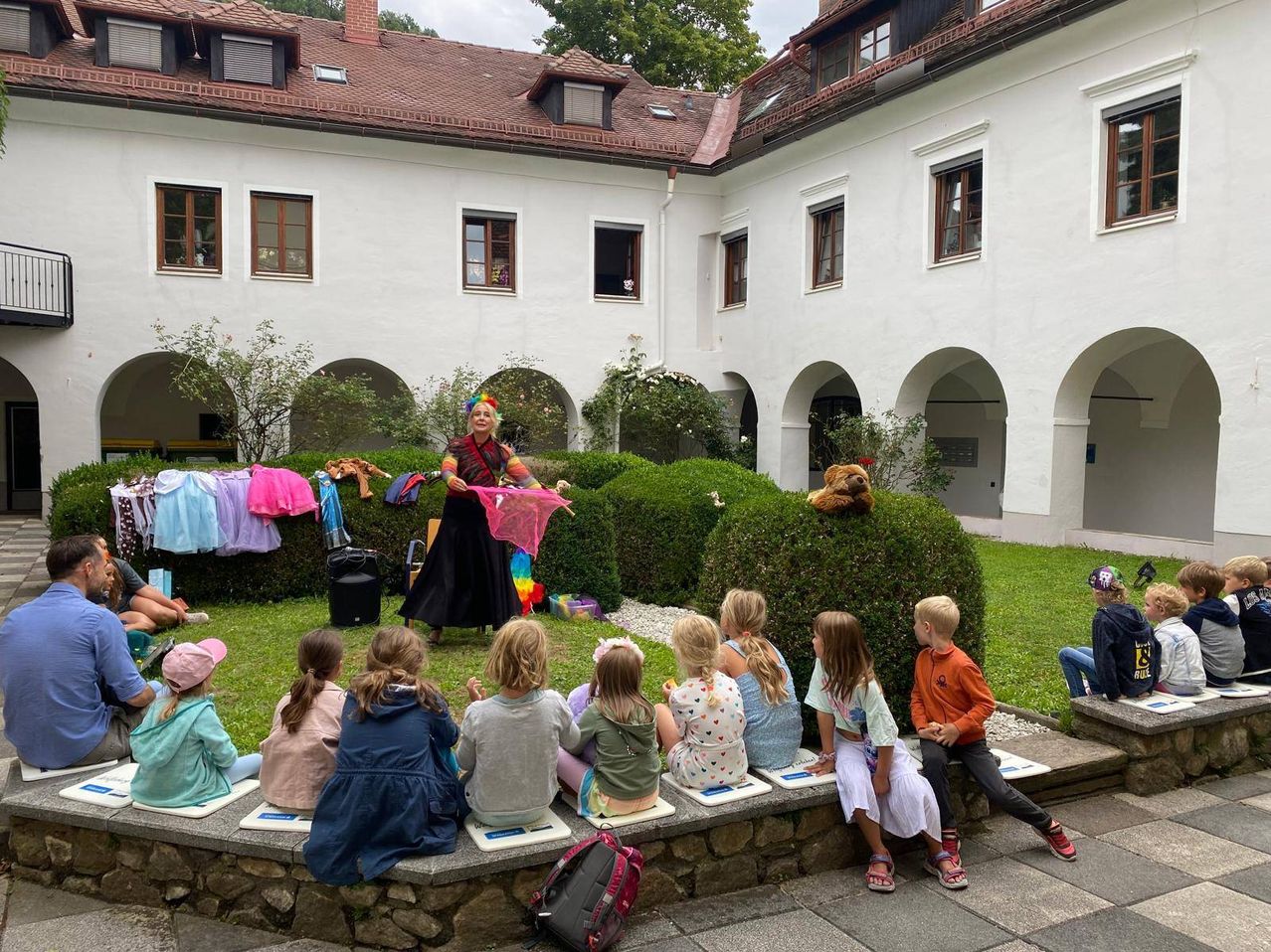 A woman in a colorful costume entertains a group of children sitting on a stone bench in a courtyard.