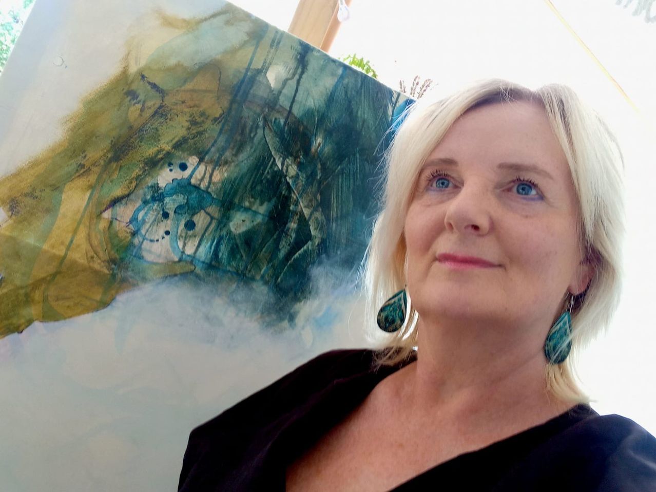A woman with blonde hair stands in front of an abstract painting, smiling at the camera. The painting behind her features blue, green, and yellow colors.