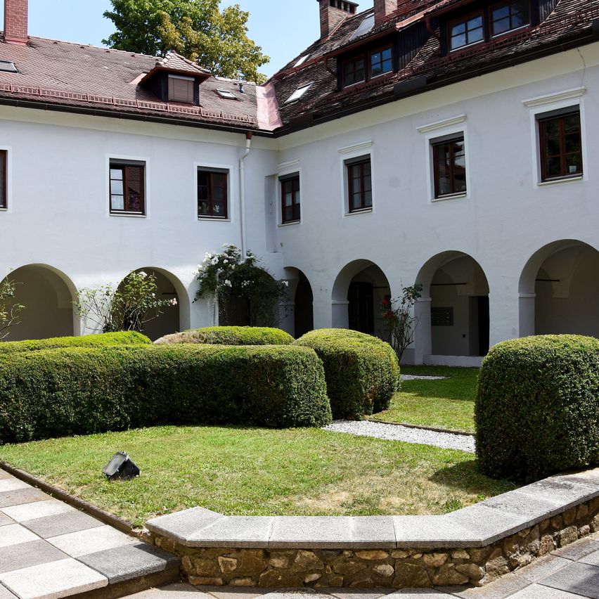 A white building with arched windows and a red roof, surrounded by a courtyard with neatly trimmed hedges and a small garden.
