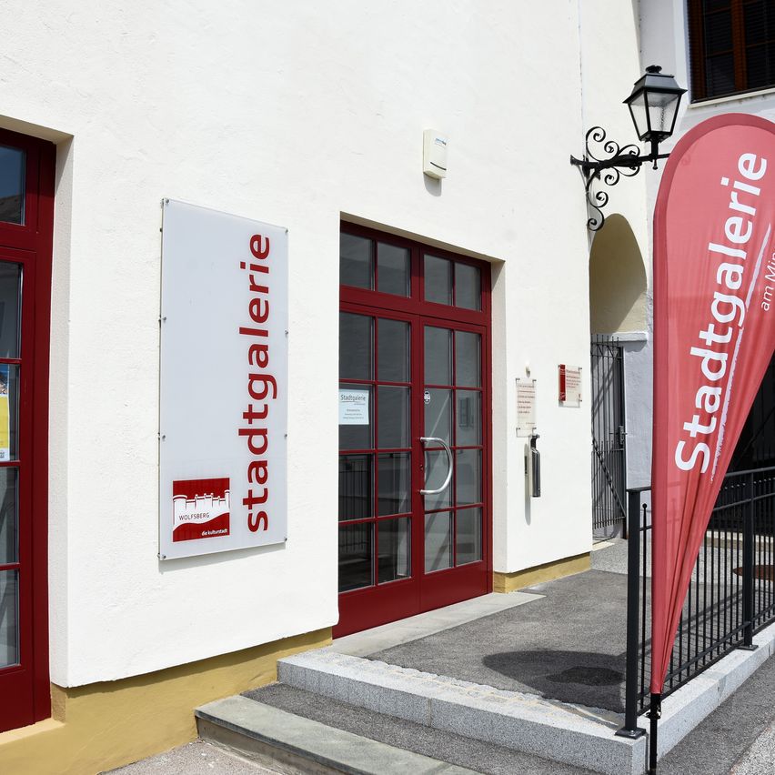 The entrance of a white building with red doors and a red banner that reads 'Stadtgalerie'. The building has a sign on the wall with the same text.