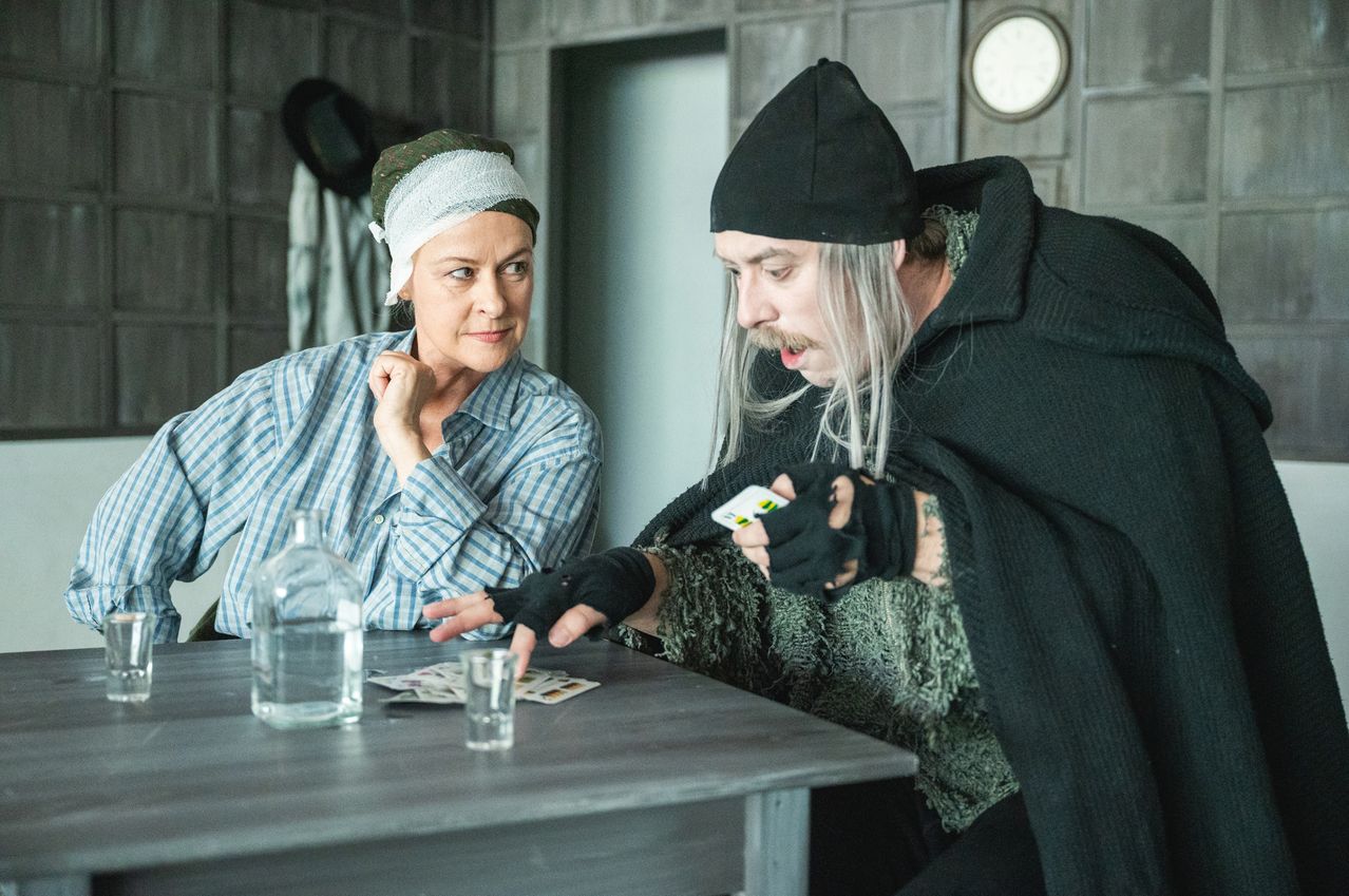 An adult woman with a bandage on her head and an adult man with long hair and a black hat are sitting at a table playing a card game.